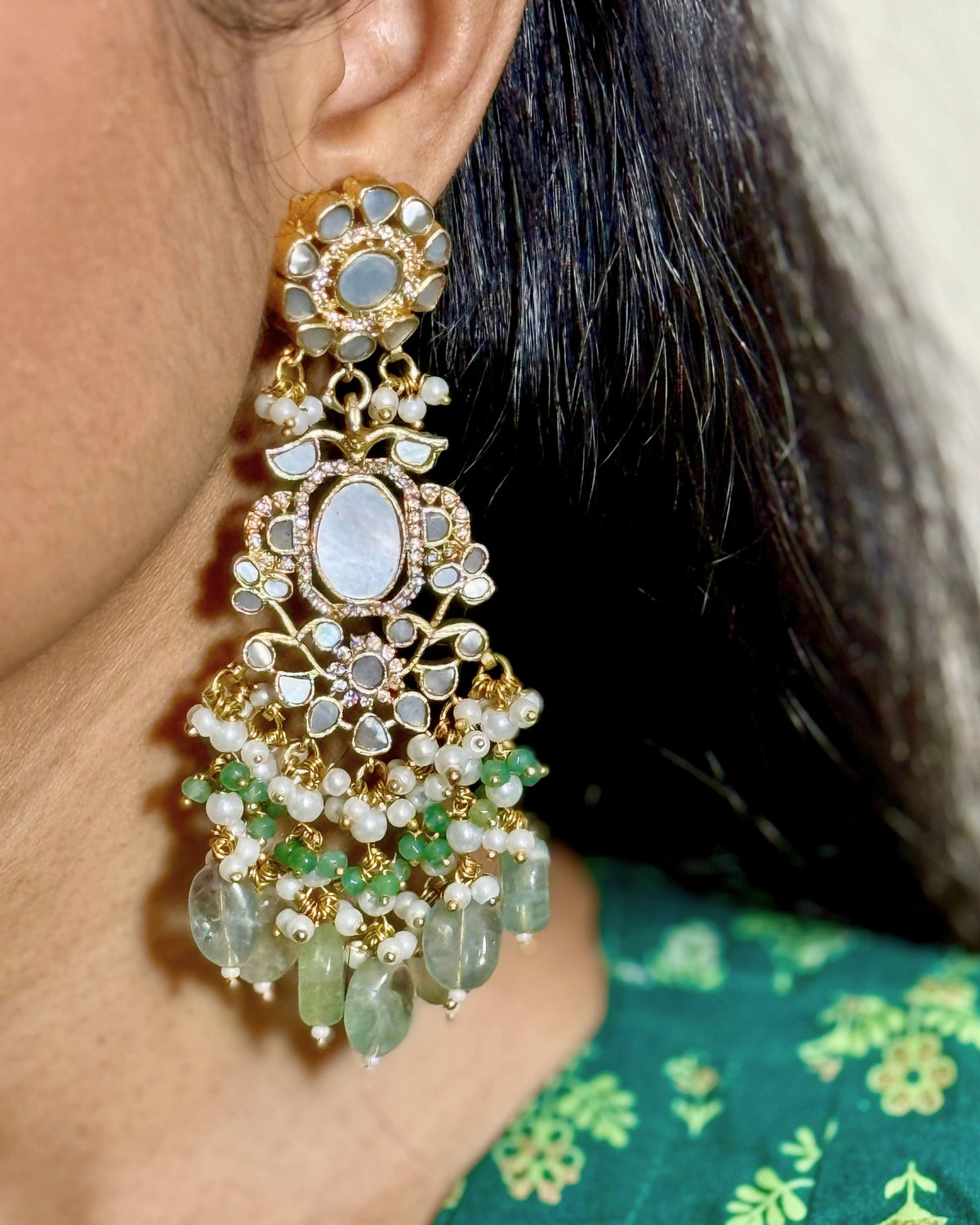 A close-up of a model wearing a Medium gold-toned dangle earring decorated with white MOP, aqua beads, and dangling Aqua seed drops, paired with a green patterned top.